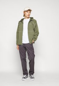Olive green zip-up jacket with a hood, white stitching, gray pants, and patterned gray sneakers. Plain white t-shirt underneath.