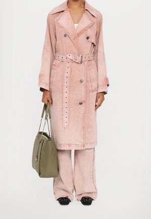 Long manteau en denim rose avec des boutons croisés et une taille ceinturée, associé à un pantalon rose ample et un sac à épaule vert.