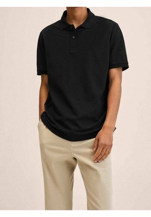 Mężczyzna w czarnej koszulce polo z krótkim rękawem i guzikami, beżowych spodniach, z jedną ręką w kieszeni, stojący na jednolitym tle.