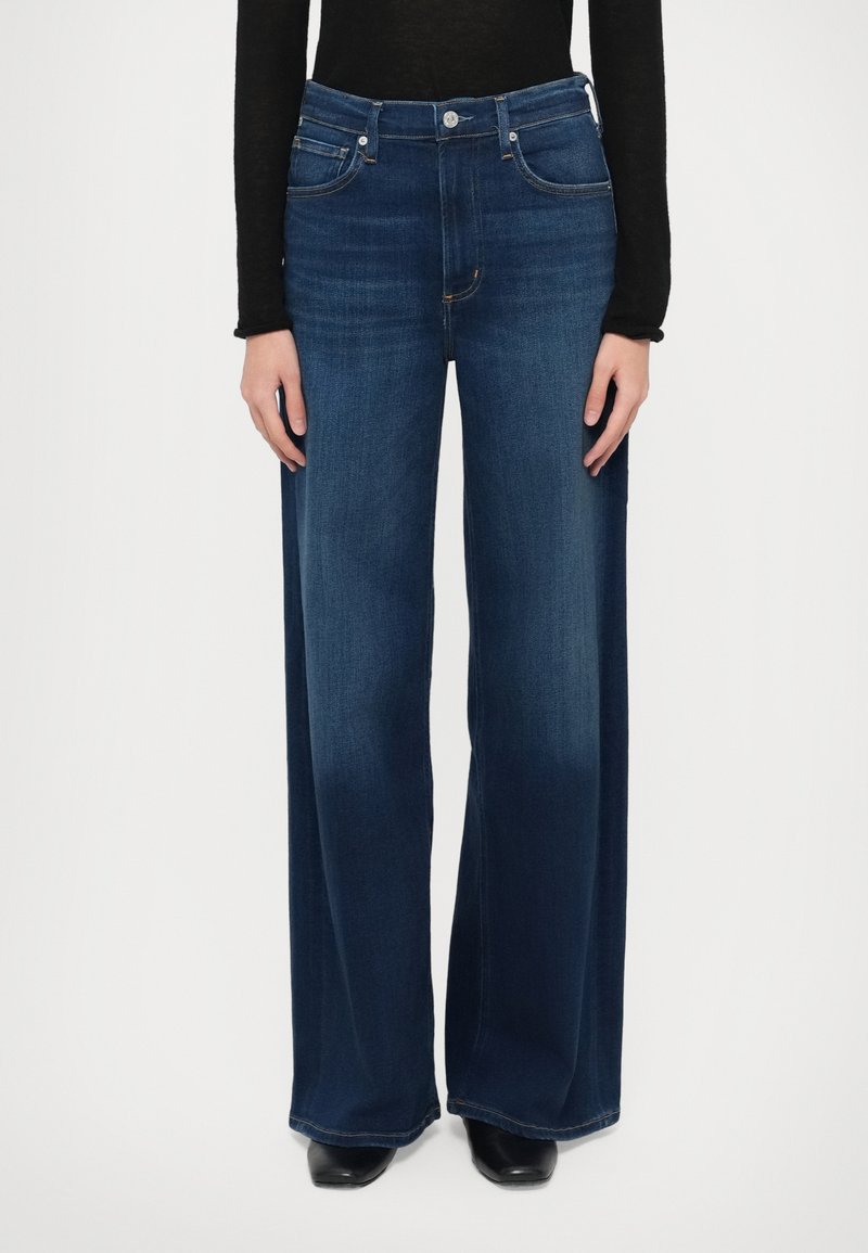 Jeans z širokimi hlačnicami v temno modrem jeansu. Imajo visok pas, pet žepov, zapenjanje na gumbe in zlato vezenje kot poudarke. Gladek tekstura.