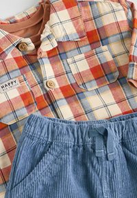 Rutig flannel skjorte med frontlommer, "Happy Big Smiles" patch, og blå corduroy bukser med elastisk talje og snor.