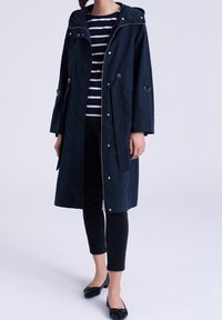 Parka imperméable marine avec capuche, fermeture éclair argentée et boutons-pression. Présente une coupe décontractée, des poches latérales et un détail de fermeture éclair sur le devant.