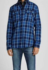 Niebieska koszula w kratę z ciemnonawigacyjnym i jasnoniebieskim wzorem, dwoma kieszeniami na piersiach, guziki na przodzie, długie rękawy i teksturowany materiał.