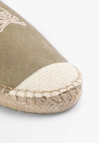 Olivenfarvet kanvassandale med en cremefarvet vævet tåhætte. Har en naturlig jutesål med syet kant. Glat tekstur og rund form.