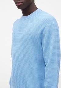 Personne portant un pull en maille texturée bleu clair à col rond, montré du cou jusqu'au milieu du torse sur fond blanc.