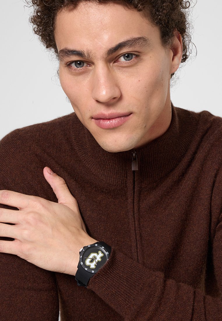 Joven con cabello rizado que lleva un suéter marrón con cremallera y un reloj de pulsera negro con una esfera gráfica audaz, posando con los brazos cruzados.