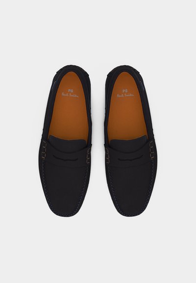 Μαύρα δερμάτινα μοκασίνια με ραμμένες λεπτομέρειες και ταμπά εσωτερικές σόλες, φωτογραφημένα από πάνω, με την ετικέτα της μάρκας να φαίνεται μέσα σε κάθε παπούτσι.