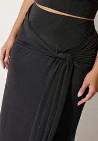 Gonna maxi nera realizzata in tessuto morbido e elasticizzato, caratterizzata da un design con frontale attorcigliato, texture liscia e dettagli di cucitura sottili.
