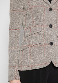 Blazer beige à chevrons avec des lignes en carreaux orange, boutons noirs, poches à rabat, et manches longues porté sur un pantalon noir.