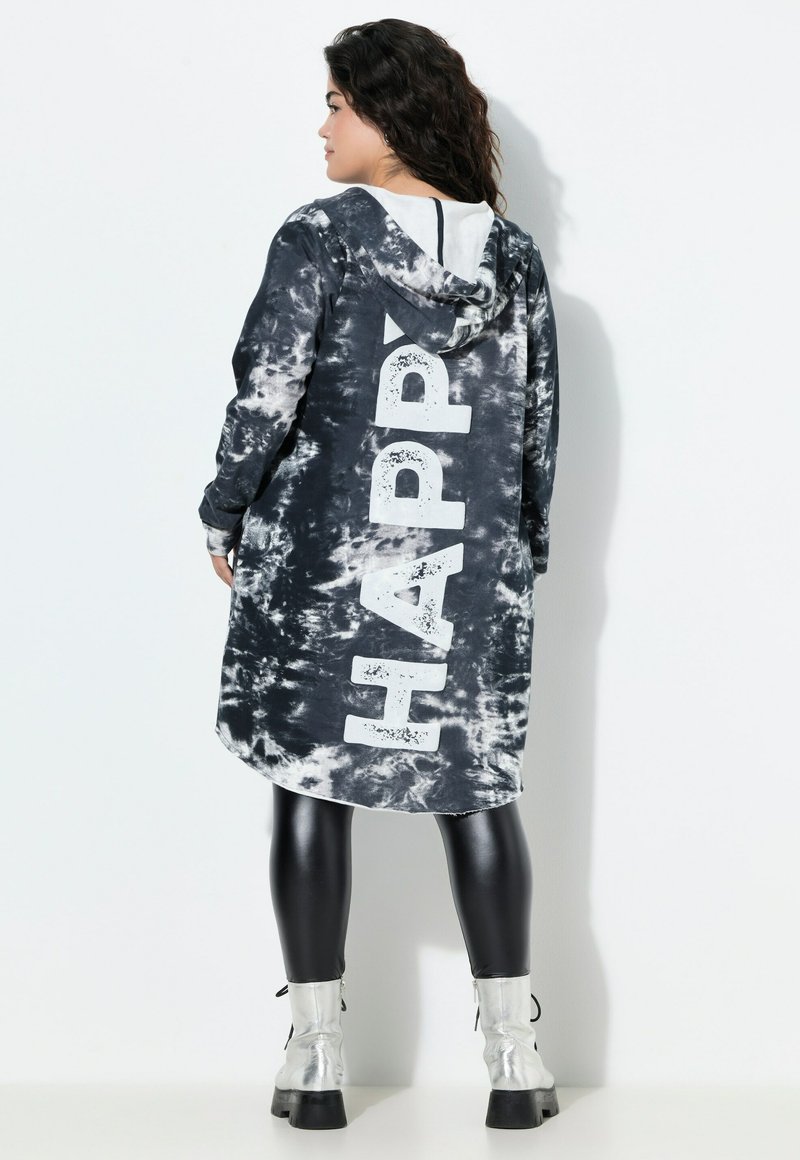 Zwart-witte tie-dye capuchonjurk met grote "HAPPY" tekst op de achterkant, voorzien van lange mouwen en een ronde onderkant, gecombineerd met glanzende zwarte leggings.