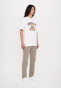 Camiseta blanca de algodón con un gráfico de osito de peluche en marrón y púrpura, combinada con pantalones beiges y zapatillas blancas.