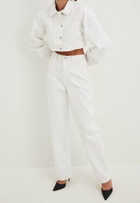 Femme portant une veste en denim blanche courte et un jean blanc taille haute, assortis à des chaussures à talons aiguilles noires, devant un fond uni.