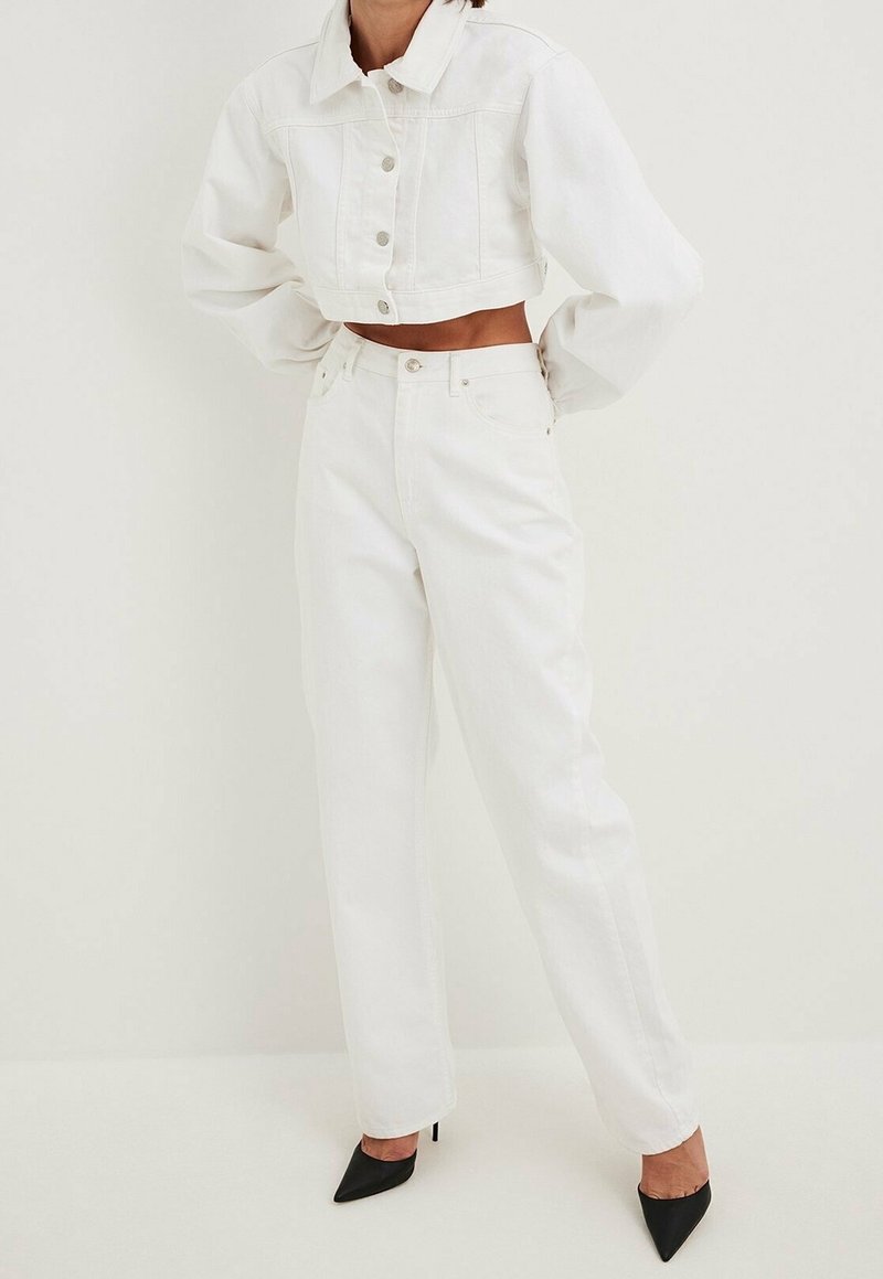 Femme portant une veste en denim blanche courte et un jean blanc taille haute, assortis à des chaussures à talons aiguilles noires, devant un fond uni.