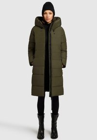 Olivgrön vadderad lång kappa med huva, dragkedjestängning, sidofickor och svarta accenter. Bärs över en svart outfit och svarta stövlar.