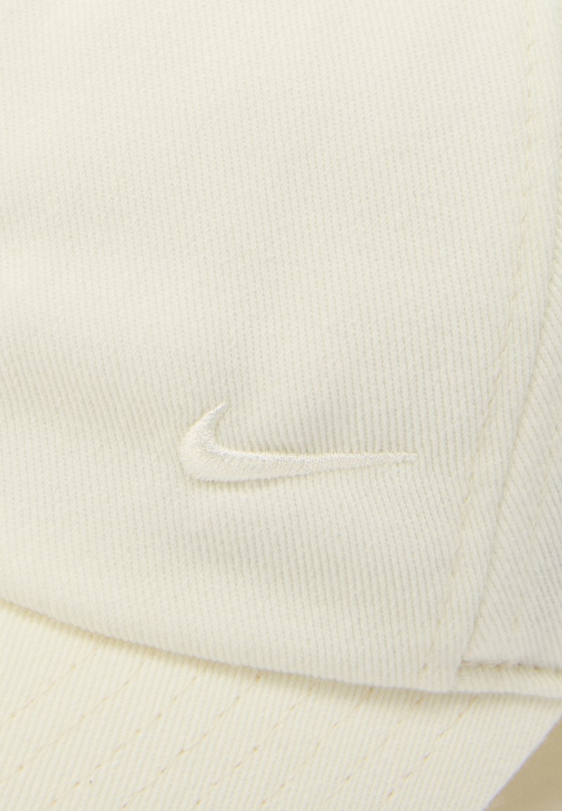 Primo piano di tessuto color avorio con un sottile logo Nike swoosh ricamato su un pannello curvo, che mostra dettagli di cuciture fini.