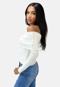 Hvid ribbet off-shoulder sweater med lange ærmer, parret med lyseblå jeans, præsenteret mod en neutral baggrund.