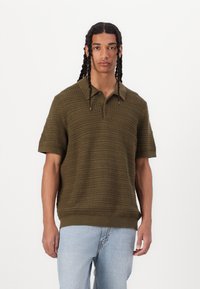 Polo shirt olive court à manches courtes avec un motif en maille texturé et un col. Ourlets et poignets côtelés, porté avec un jean bleu clair.