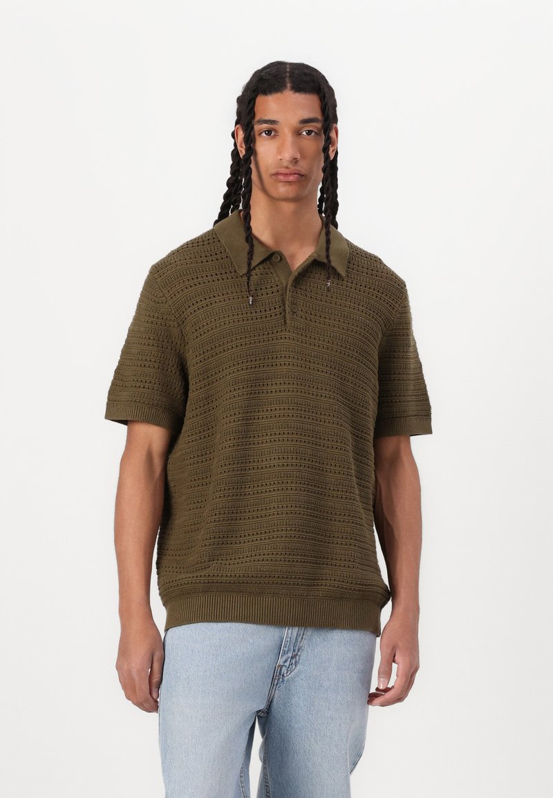 Polo shirt olive court à manches courtes avec un motif en maille texturé et un col. Ourlets et poignets côtelés, porté avec un jean bleu clair.