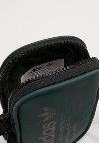 Zelená zipová taška Adidas s texturovaným povrchem, černým zipem a logem vytištěným na přední straně; obsahuje látkovou vnitřní podšívku.