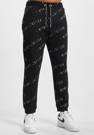 Sorte joggingbukser med "Carlo Colucci"-tryk over det hele, elastisk linning med snore, båret med hvide Nike sneakers.