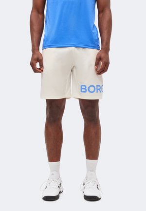 Homme portant un short crème avec le texte bleu "BORG" sur la jambe gauche, des chaussures de sport blanches, des chaussettes blanches et une chemise bleue.
