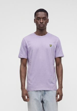 Mand iført en lyse lilla kortærmet t-shirt med en lille gul fuglelogo på venstre bryst og lyseblå jeans, står mod en ensfarvet baggrund.