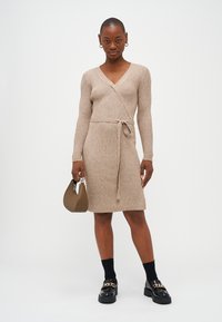 Beige ribbestrikket kjole med omslagsdesign, lange ermer og knyttet midje. Kombinert med svarte klumpete sko og en liten brun veske.