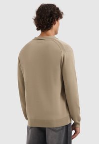 Mann mit lockigem Haar, der einen beigen Langarmpullover und dunkelgraue Hosen trägt, von hinten vor einem schlichten Hintergrund betrachtet.