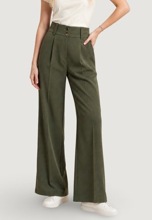 Femme portant un pantalon taille haute, jambes larges, vert olive, avec deux boutons et des baskets blanches, debout avec une main dans la poche.