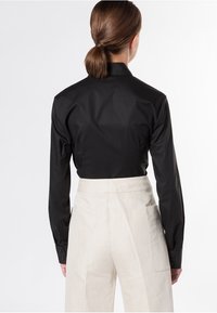 Persona con capelli castani raccolti in una coda di cavallo bassa, che indossa una maglietta nera a maniche lunghe infilata in pantaloni beiges a vita alta con tasche laterali, vista di spalle.