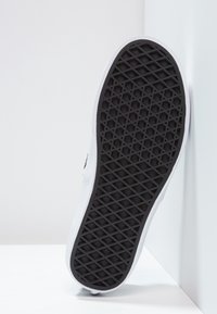 Bílé tenisky s texturovanou černou gumovou podešví s diamantovým vzorem. Obuv má čistý design a zakulacené okraje.