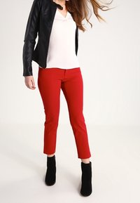 Donna che indossa una giacca di pelle nera, una maglietta bianca a V, pantaloni rossi slim-fit e stivaletti neri, in piedi contro uno sfondo bianco uniforme.