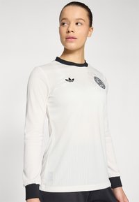 White long-sleeve athletic shirt with black ribbed collar and cuffs, featuring a small black logo on the chest and vertical ribbed texture.
