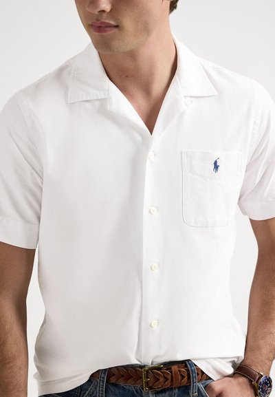 Balta īsroku krekli ar pogām, izgatavota no kokvilnas, ar izgriezumu apkakli, kabatu ar zilu logotipu un pogu aizdari. Redzams brūns austs josta.