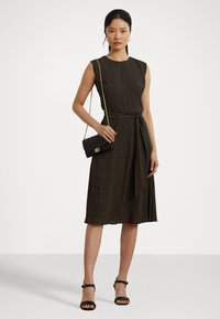 Black sleeveless dress with a fitted bodice and A-line skirt, featuring a yellow and white polka dot pattern, paired with a small black bag.