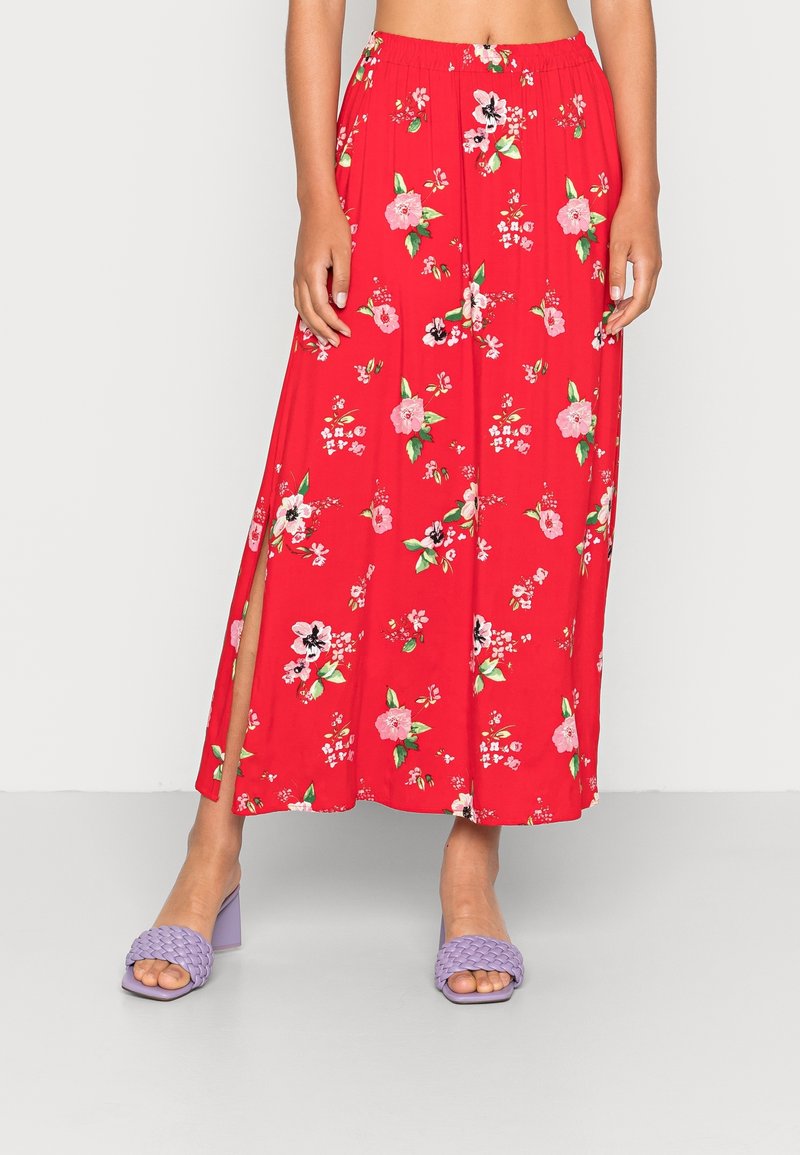 Jupe longue rouge à fleurs avec fente latérale portée avec des sandales à talons carrés tressées lavande à bout ouvert sur une personne debout.