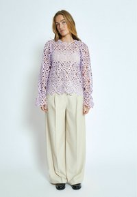 Top di pizzo viola con intricati motivi floreali e maniche lunghe, abbinato a pantaloni beige a gamba larga e scarpe nere.