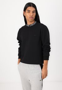 Hanorac negru cu guler înalt, având un logo alb "Lacoste", confecționat dintr-un material neted, asortat cu pantaloni de trening gri.