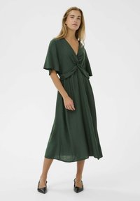 Rochie maxi verde cu un front rasucit, maneci scurte și largi, și o siluetă fluida. Purtați cu tocuri negre cu vârful ascuțit.