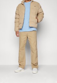 Beżowa kurtka puchowa z sztruksu na zamek, zestawiona z jasnoniebieskim t-shirtem i beżowymi spodniami oraz białymi trampkami. Na kurtce widoczne jest małe logo.