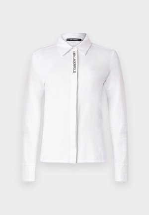 Chemise blanche à manches longues en coton avec un col, présentant un logo noir vertical sur le devant et des poignets boutonnés.
