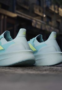 Kerge sinise ja kahvatu kollase värvusega Adidas Ultraboost jooksujalatsite paar, tagantpoolt nähtav, asetatud karedale välispinnale.