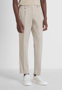Pantaloni in lino beige con una vestibilità sartoriale, caratterizzati da pieghe frontali e chiusura con bottone, abbinati a sneakers bianche per un look casual.