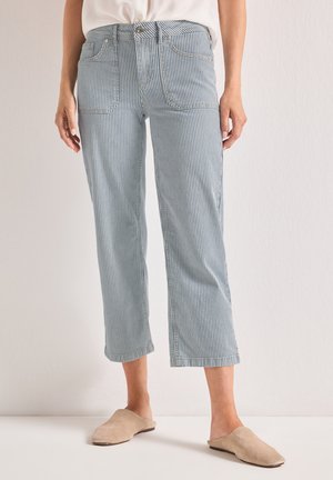 Kvinde iført kortede blå og hvide lodrette striped bukser og beige slip-on sko, der står mod en ensfarvet baggrund.