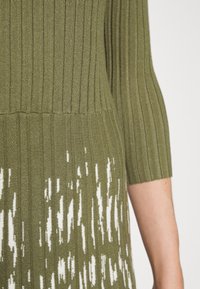 Robe en maille présentant un corsage côtelé vert olive avec un motif abstrait blanc sur la jupe. Manches courtes et tissu texturé.