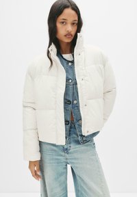 Hvid pufferjakke med høj krave og trykknaplukninger, båret over et denim-outfit. Har quiltet design og en let cropped længde.