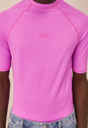 Nahaufnahme einer Person, die ein leuchtend pinkes kurzärmeliges Shirt mit dem "GCDS"-Logo auf der Brust und hellblaue Jeans trägt.