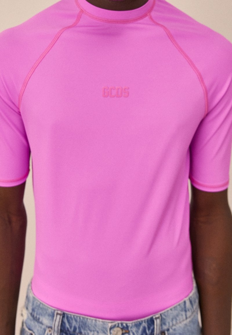 Gros plan d'une personne portant un t-shirt à manches courtes rose vif avec le logo "GCDS" sur la poitrine et un jean en denim bleu clair.