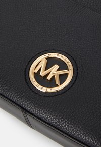 Černá kožená taška se zakulaceným zlatým emblémem s nápisem "MK" a "MICHAEL KORS." Texturovaný povrch se šitými detaily.