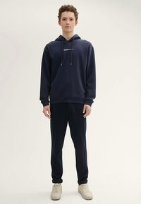 Felpa con cappuccio blu navy con coulisse e logo, abbinata a pantaloni blu navy stretti e sneakers beige su sfondo chiaro.